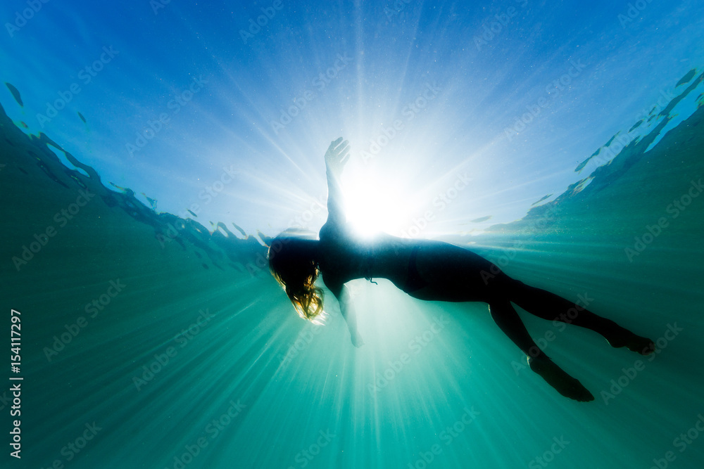 A beautiful woman floats on her back in the ocean as she is surrounded be bright ethereal light and sun rays.